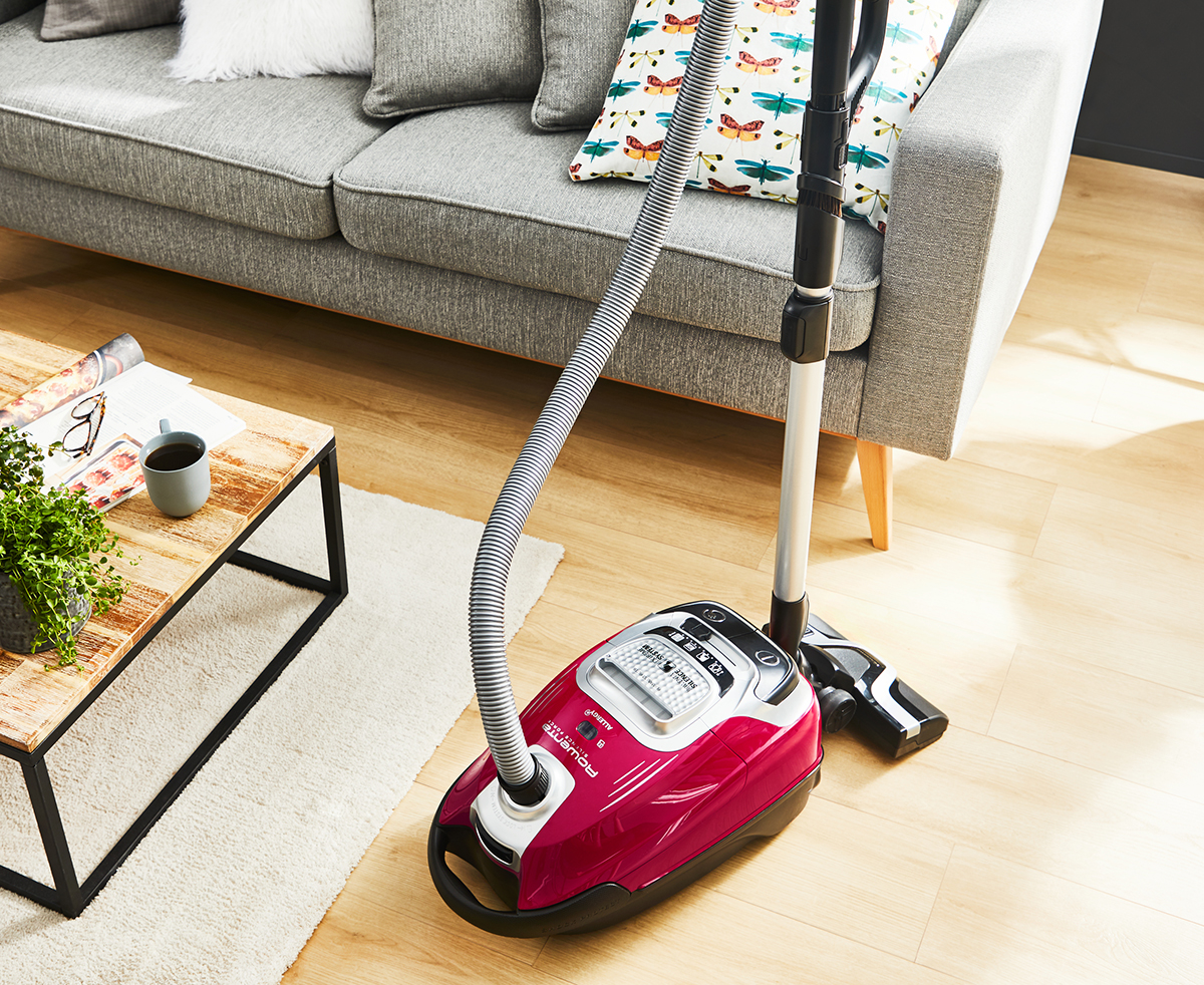 A pink vacuum cleaner is positioned near a gray sofa and a wooden coffee table with a plant and a cup. Sunlight illuminates the scene.
