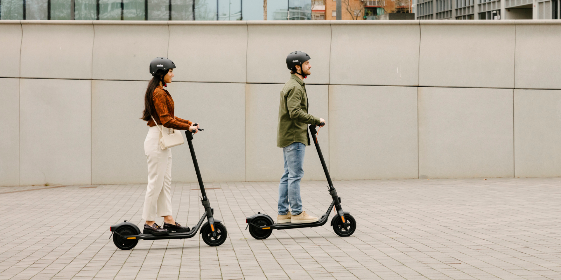 A man and a woman ride electric scooters side by side on a paved area, both wearing helmets.