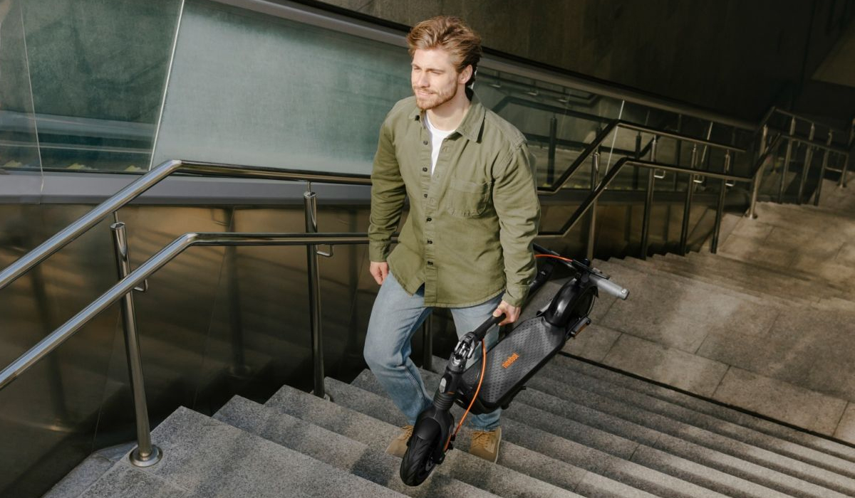A man is walking up stairs while carrying an electric scooter, dressed in a green shirt and jeans.