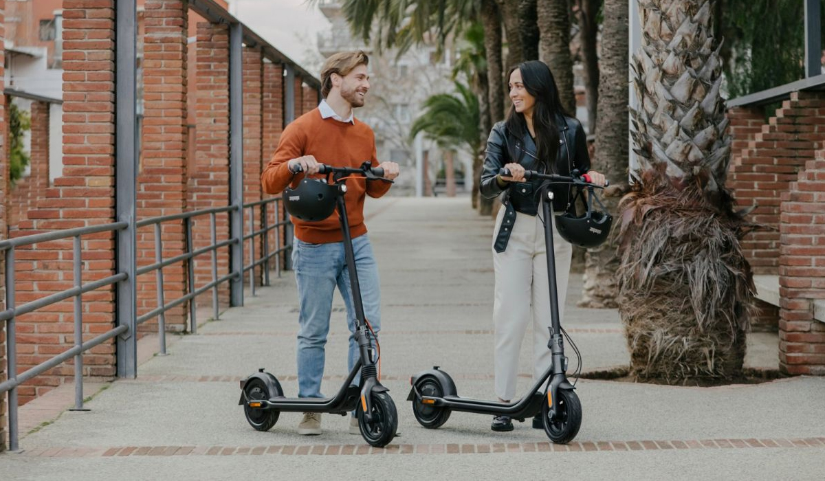 A man and woman stand on a pathway, each holding an electric scooter and helmet, smiling at each other. Palm trees line the background.
